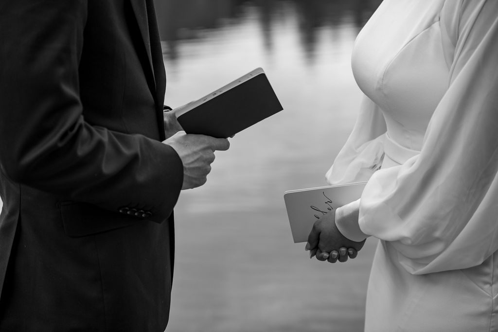 A man and woman holding vow booklets while saying their vows outdoors. The woman wearing a wedding dress with sleeves. The man is in a suit. The image is in black and white.