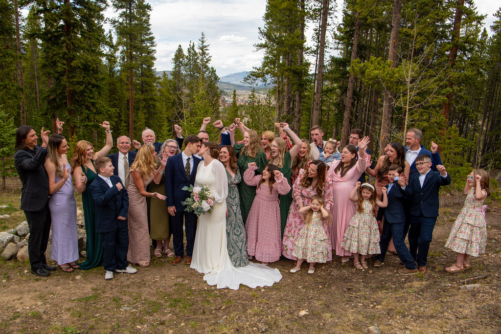 A group of people standing outdoors, posing for a photo in front of a tree. The scene captures a wedding celebration, featuring individuals in formal attire, including a bride in a beautiful wedding dress and the bridal party dressed elegantly. The sky is visible above them, enhancing the joyful atmosphere of the marriage ceremony.