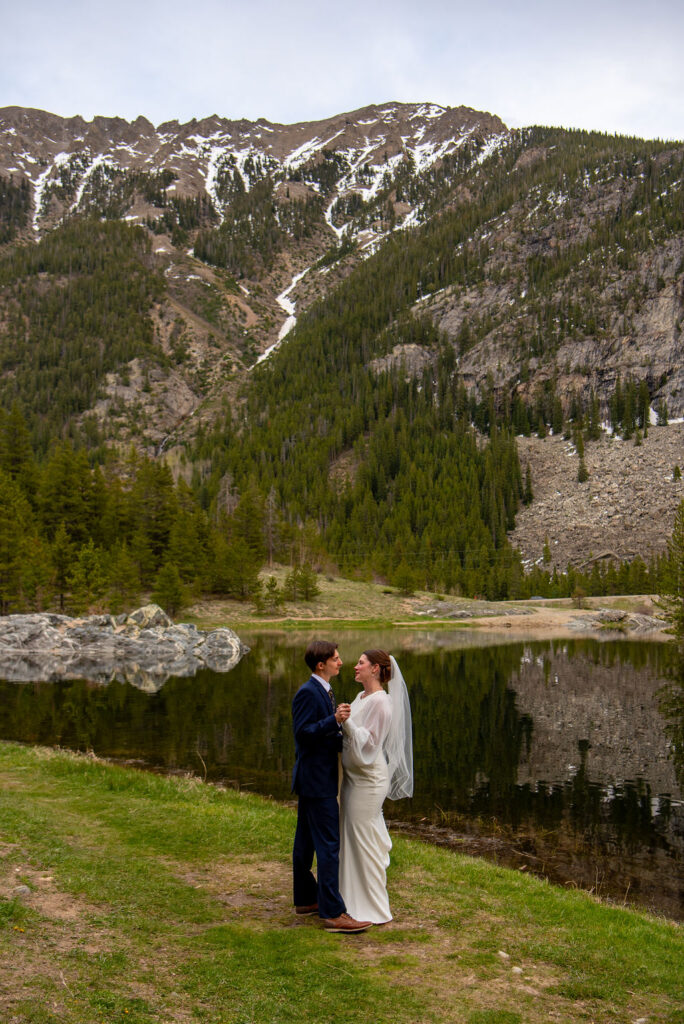 A bride and groom stand together in front of a serene lake, surrounded by lush grass and mountains under a clear sky with clouds. The woman is dressed in a wedding gown while the man wears formal attire. The Breckenridge wedding photographer captures this moment of the couple dancing on the grass
