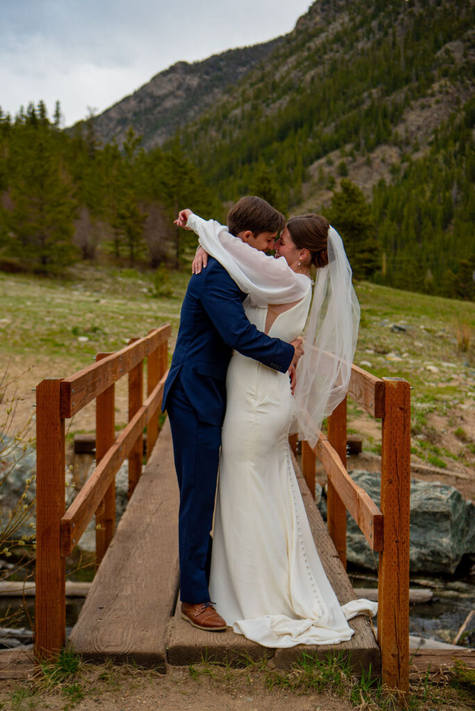 Breckenridge wedding photographer captures a moment between a bride and groom kissing on a bridge, with a scenic background of grass and mountains under a clear sky. The woman is wearing a wedding dress.