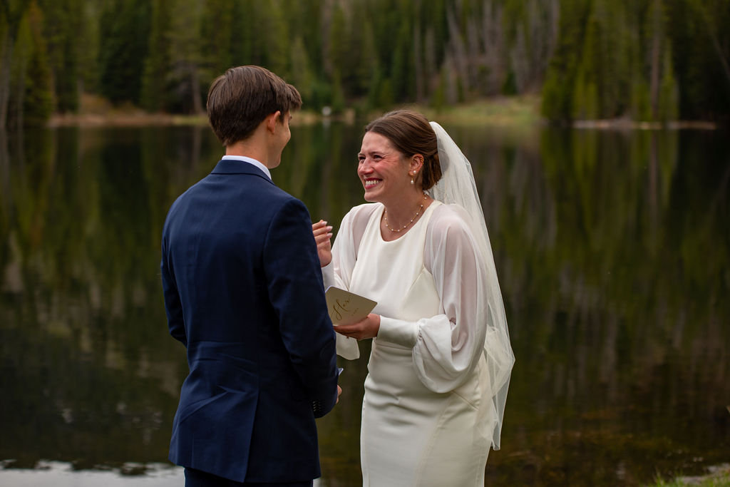 A man and woman standing facing each other while they exchange wedding vows. The woman wearing a wedding dress and smiling at the groom. They are near a lake surrounded by trees in the Colorado mountains.