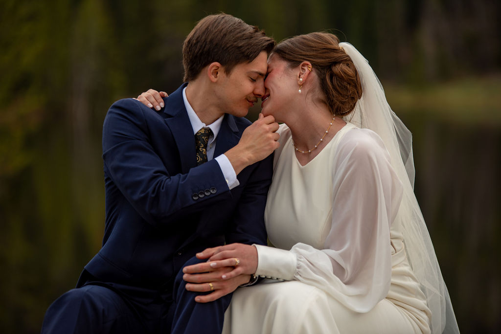 A man in a suit and a woman in a wedding dress sitting on a bench, embracing each other with affection outdoors. The Breckenridge wedding photographer captures the moment before the couple goes in for a kiss.