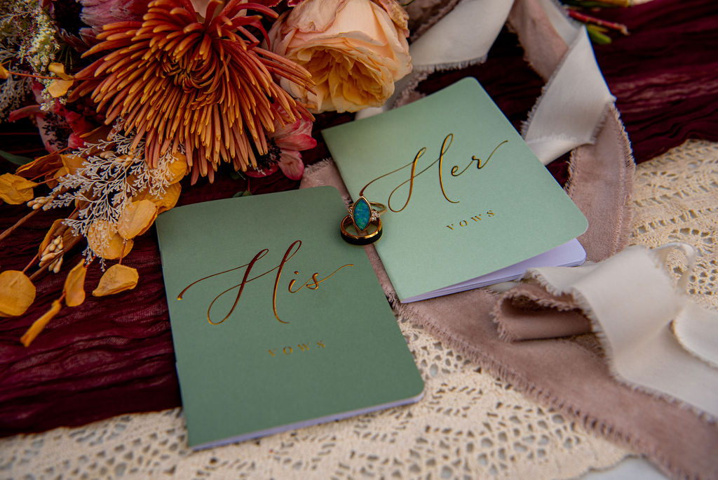 Two green vow booklets labeled "his" and "hers" are arranged next to a bouquet of flowers indoors with their wedding rings placed between the books. The Colorado wedding photographer captures the flatlay to remember the wedding details.