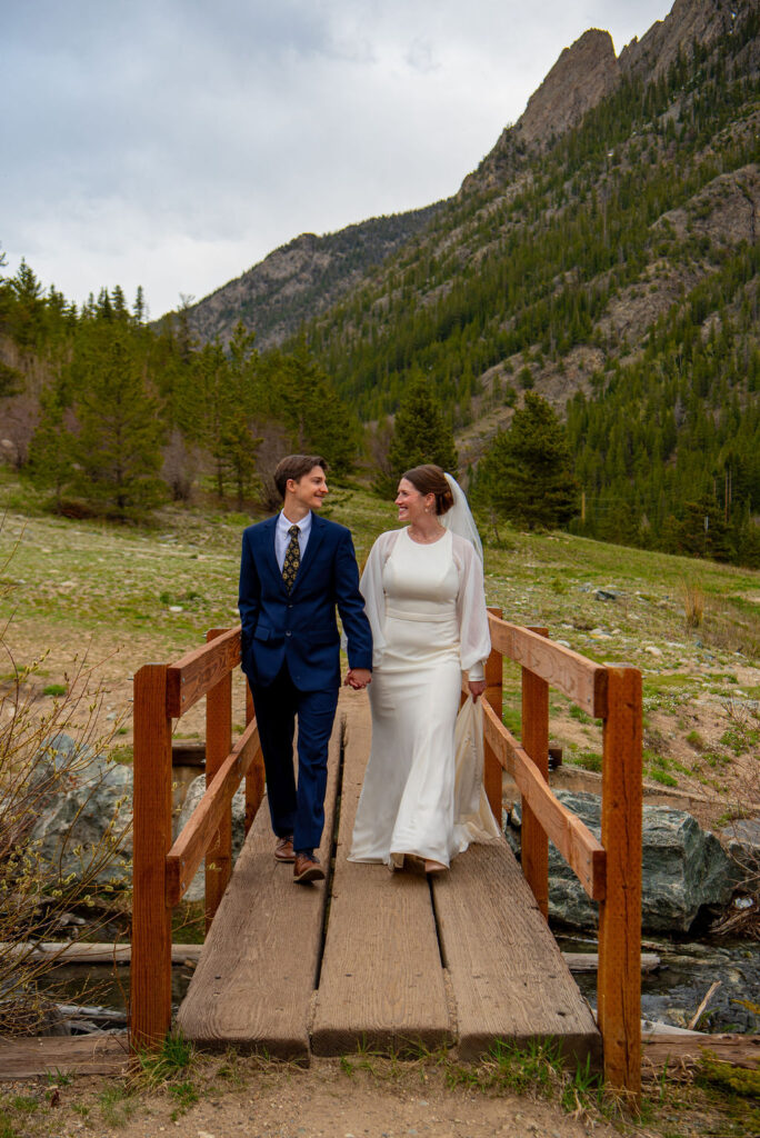 A bride and groom walk hand in hand across a wooden bridge after exchanging wedding vows, surrounded by lush green mountains of Colorado and trees, celebrating their wedding day.
