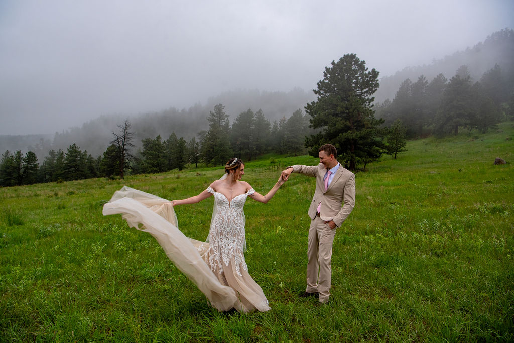 In a misty field, a bride and groom walk hand in hand. The groom gives the bride a twirl and she shows off her dress. This moment showcasing the beauty of Colorado weddings and the Colorado wedding photographer skill.