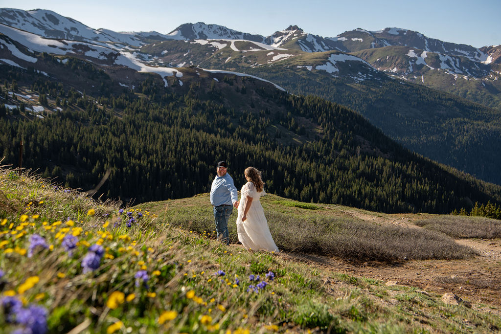 The Colorado wedding photographer captures a moment of a couple stands hand in hand on a grassy hill, overlooking a stunning mountain landscape with snow-capped peaks and wildflowers in bloom.