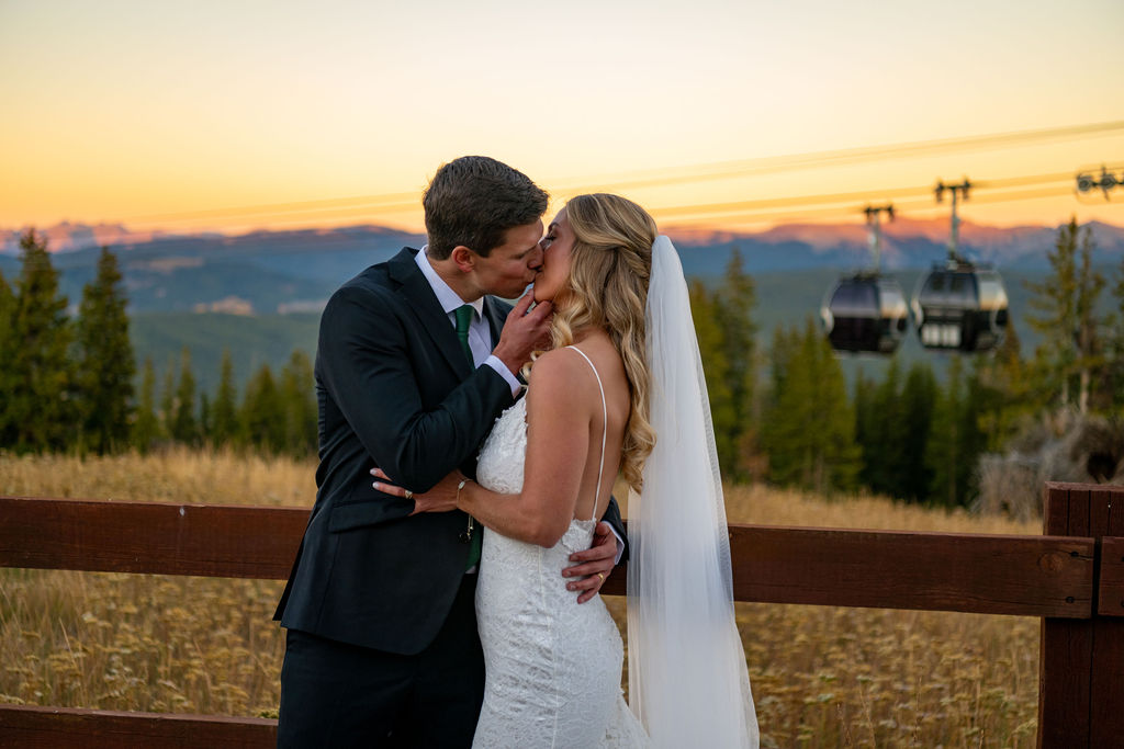 A couple shares a romantic kiss at sunset, surrounded by the Colorado mountains and trees, with a ski lift running in the background.