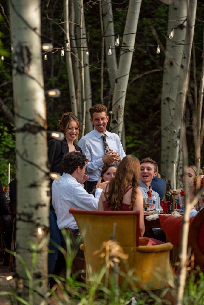 The Colorado wedding photographer captures a lively outdoor dinner party surrounded by tall trees and string lights, with guests engaged in conversation around a beautifully set table.
