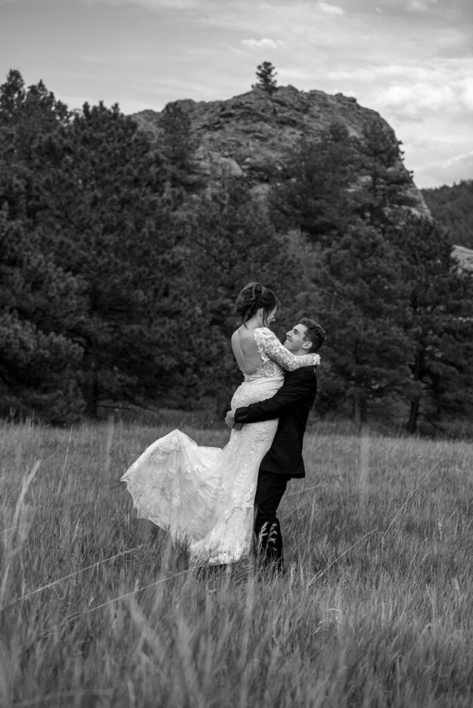 A bride in a lace gown is lifted by a groom in a black suit, surrounded by tall grass and evergreen trees against a rocky backdrop.