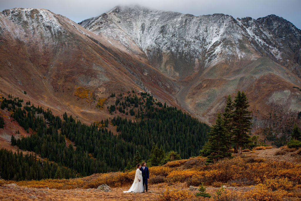 A couple standing together in front of a majestic mountain range with snow capped peaks, surrounded by lush greenery and yellow foliage on the ground. The terrain showcases the rugged features of the mountain and jagged ridges at the turn of fall.