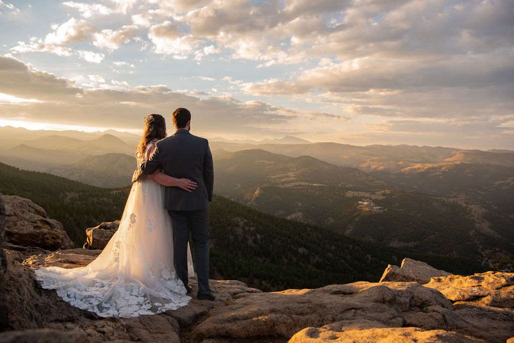 A newlywed couple stands on a rocky outcrop, embracing with a breathtaking mountain sunset in the background.