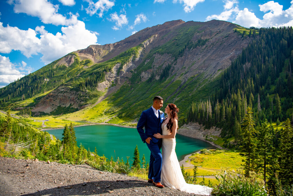 Not sure where to elope in Colorado, A couple in formal attire stands by a serene turquoise lake, surrounded by lush green mountains and a bright blue sky with clouds showing why this is the best elopement location.