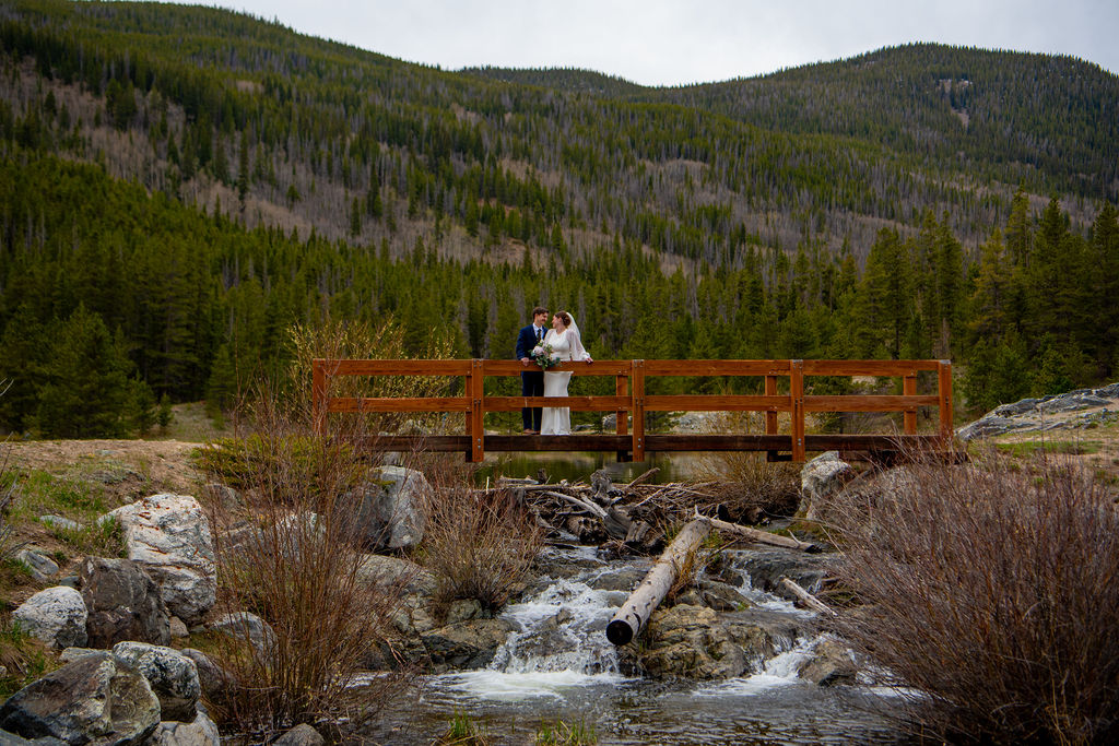 A bride and groom stand on a wooden bridge over a stream, surrounded by lush mountains and greenery, capturing a romantic moment and showing where to elope in Colorado if a couple wants adventure!