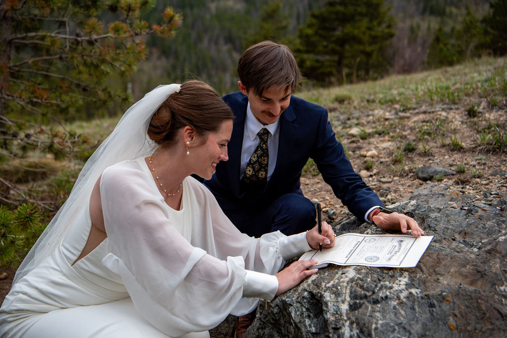 A man in a suit and a woman in a wedding dress are signing a wedding document outdoors, with grass underfoot and signing on a rock.