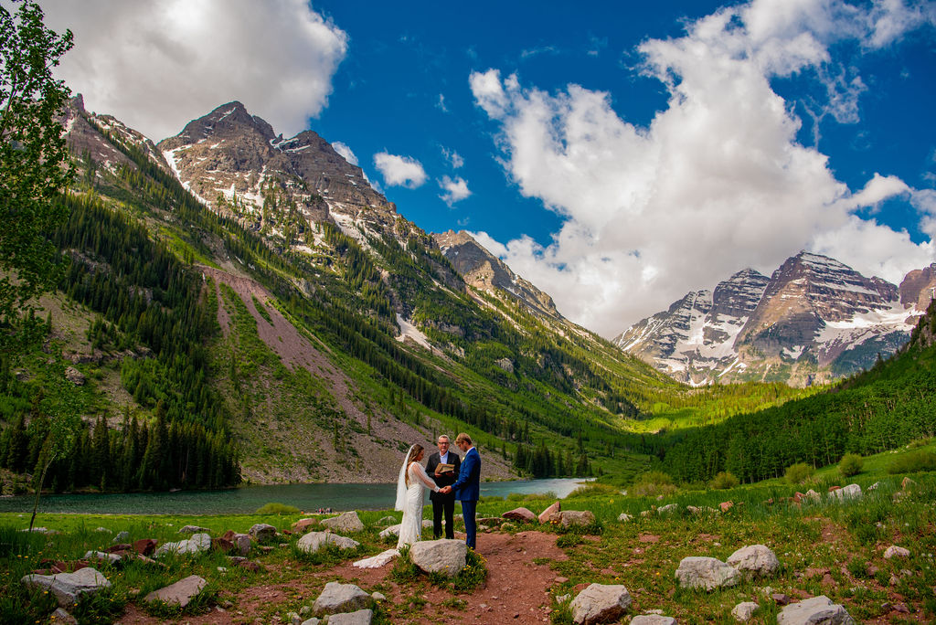 A couple exchanges vows in a scenic mountain setting, with lush greenery and snow-capped peaks under a bright, partly cloudy sky. The setting is in Aspen Colorado at the Maroon Bells. This is where to elope in Colorado if you want iconic mountain views