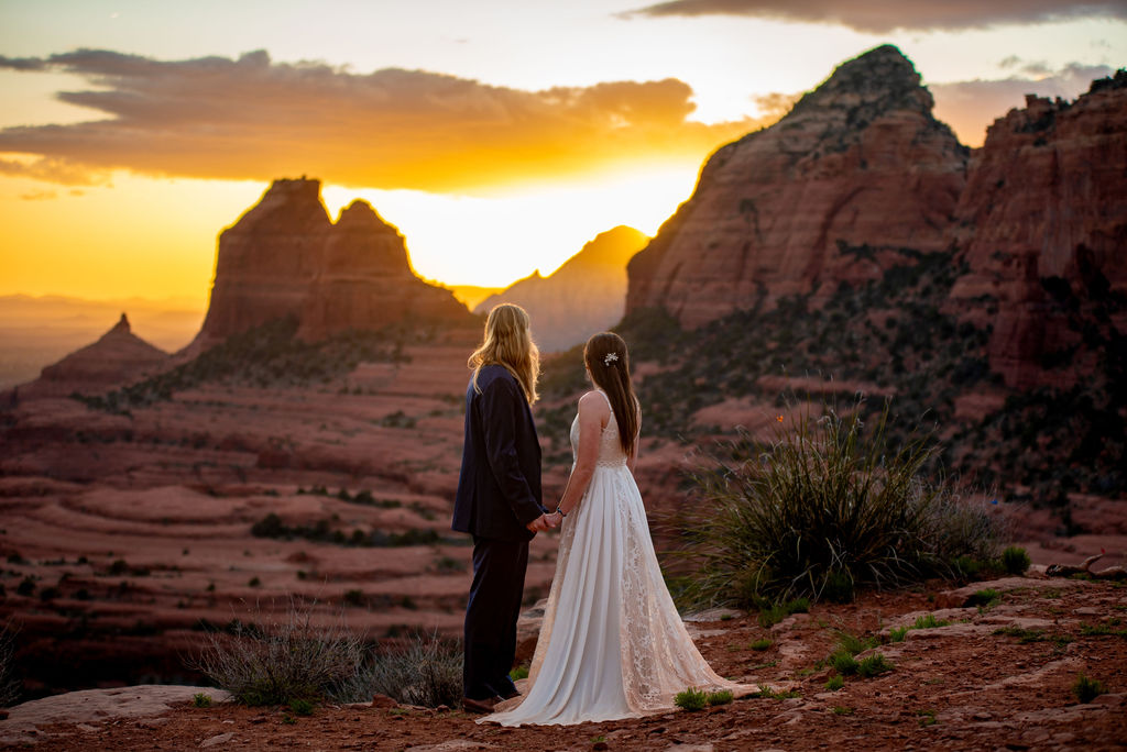 A Sedona elopement photographer captures a photo of a couple in wedding attire stands holding hands, facing a sunset over red rock formations in Sedona AZ.
