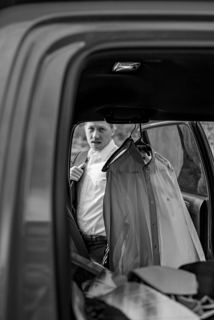 Groom stands outside a car, visible through the open door, adjusting his jacket. Several dress shirts on hangers are hanging inside the car. A quiet moment captured by a Sedona elopement photographer.